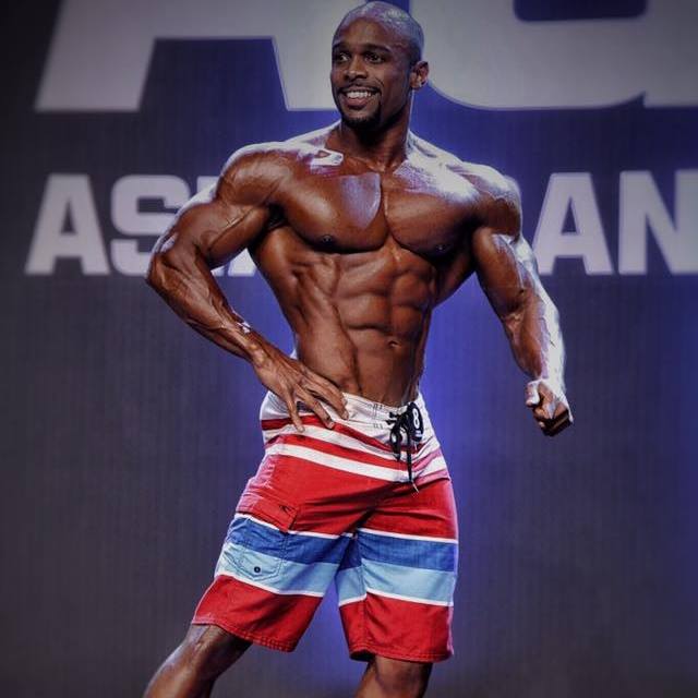 Ryan Hinton posing on the stage, showing his physique from the side, his abs, arms, and chest looking contest-ready