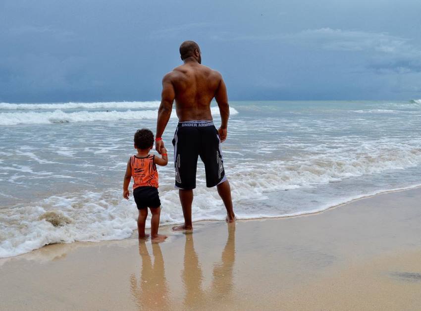 Ryan Hinton on a beach with his son on the left, Ryan's back and arms looking big and aesthetic