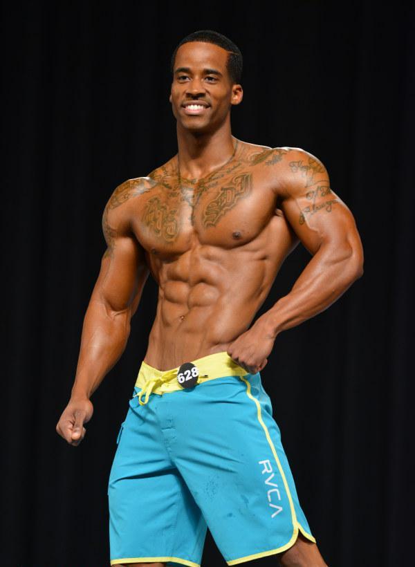 Andre Smith posing on the stage in light blue trunks, smiling at the audience, looking ripped