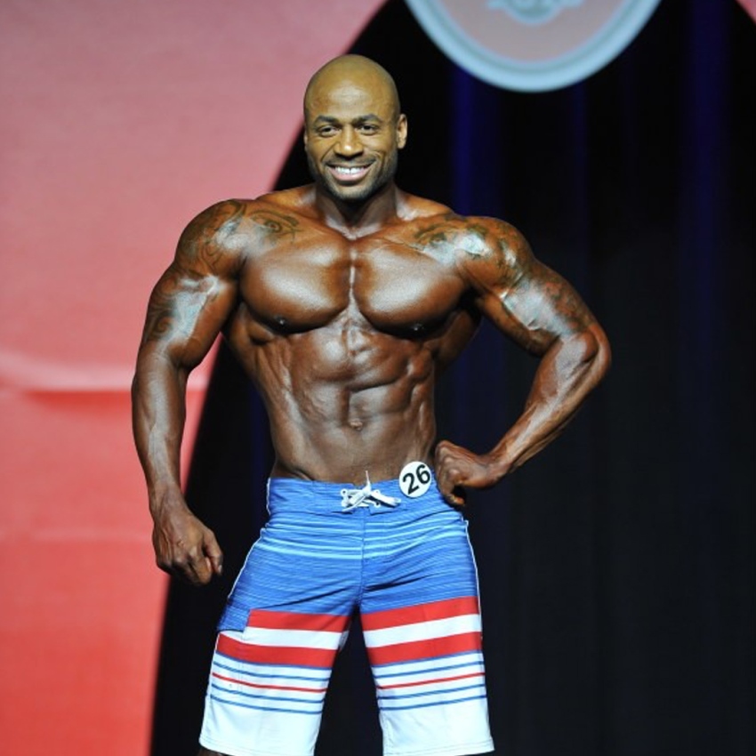 Jacques Lewis posing on the Olympia stage, smiling at the audience, and showing his ripped physique