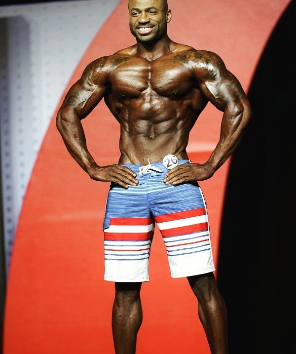 Jacques Lewis on the Mr. Olympia stage, doing a front pose and smiling at the auditorium
