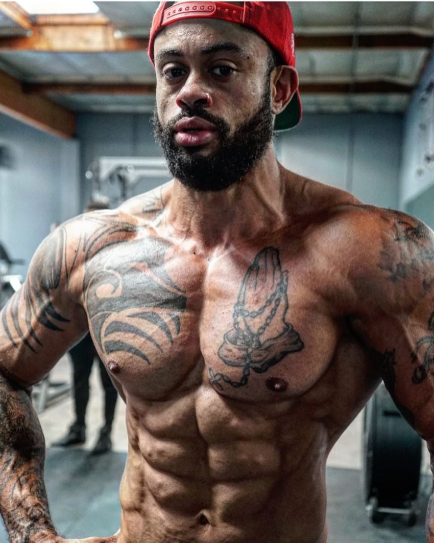 Kai Spencer flexing his ripped abs, tattoed cheest, and muscular arms as he looks directly at the camera