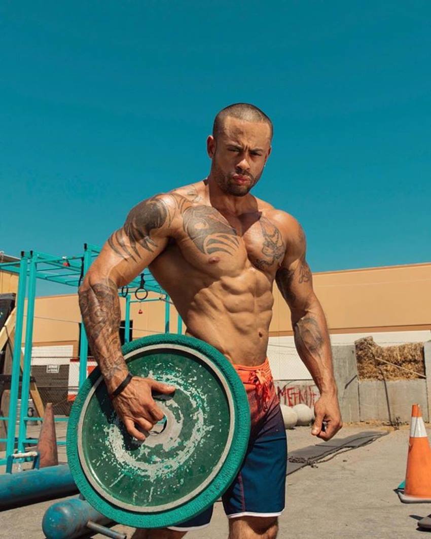 Kai Spencer holding a green weight plate, standing shirtless outdoors in the sun, looking fit and lean