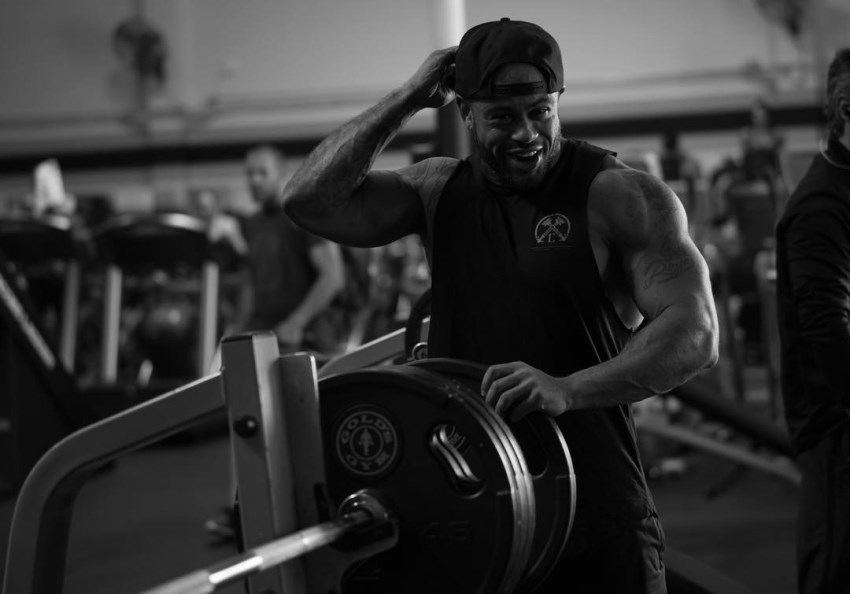 Kai Spencer putting a weight plate on a bar in the gym, wearing a sleeveless shirt, looking muscular and fit