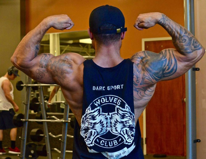 Kai Spencer doing a back double biceps pose in a sleeveless shirt, showcasting his ripped and muscular arms, shoulders, and back