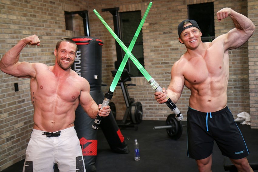 Chris Powell and Steve Cook holding green lightsabers, posing shirtless alongside each other
