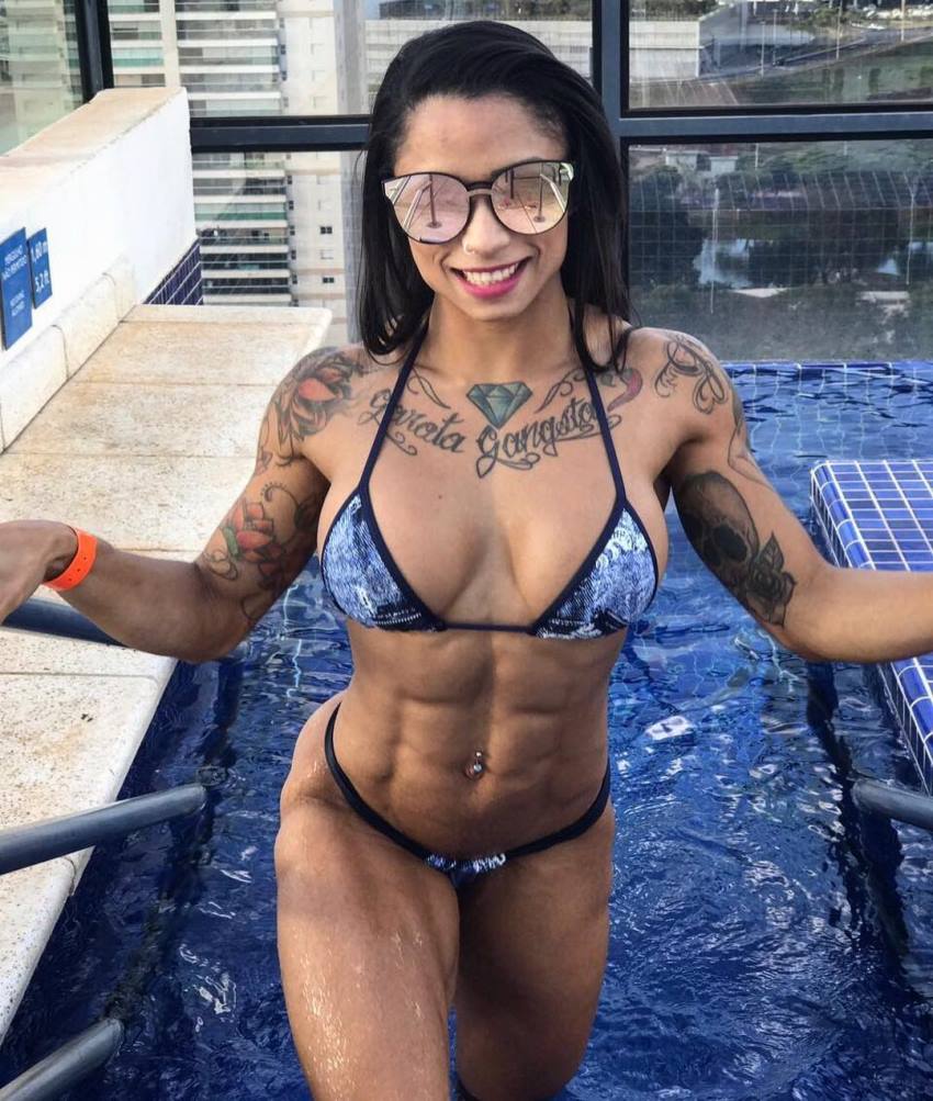 Elah Bittencourt standing in a pool and smiling at the camera, flexing her ripped abs