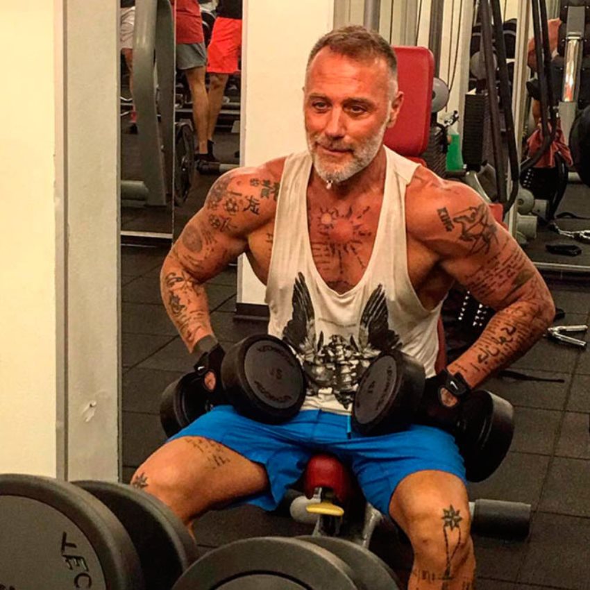 Gianluca Vacchi sitting on a bench in the gym, preparing to do an exercise with dumbbells