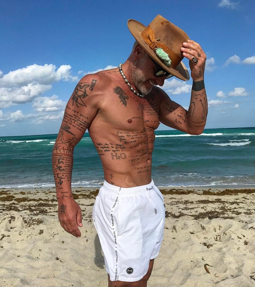 Gianluca Vacchi stainding on the beach with a hat on, looking fit