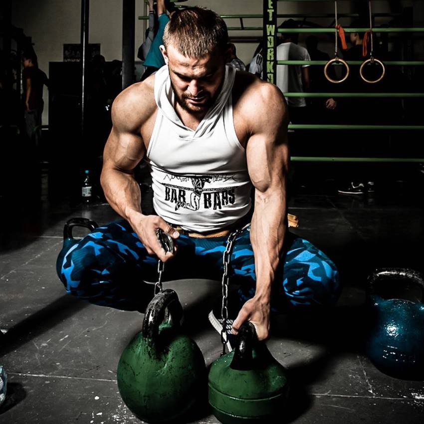 Islam Badurgov holding kettlebels on the floor, wearing a white tank top, looking muscular and ripped