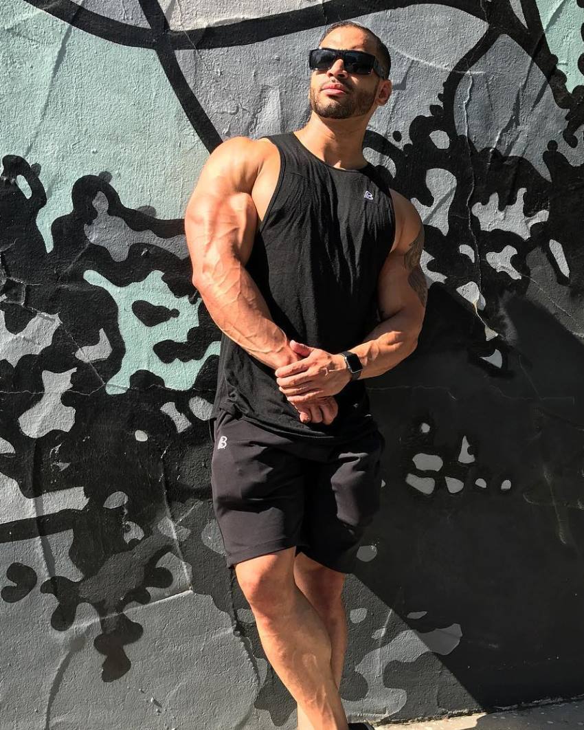 Jamie LeRoyce leaning against the wall in his black tank top, shorts, and sunglasses, his arms looking muscular and ripped