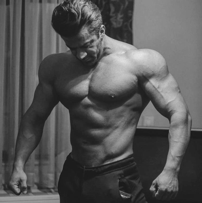 Mohammad Kashanaki posing shirtless in a room, looking massive and ripped