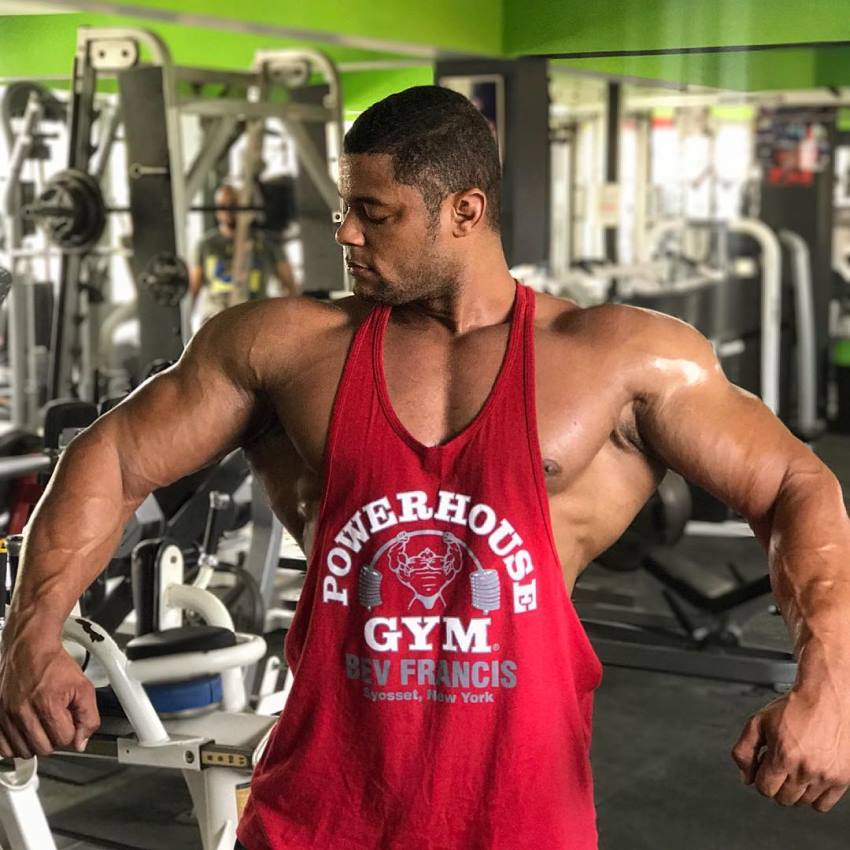 Robert Galva standing in a gym in a red tank top, spreading his arms and lats