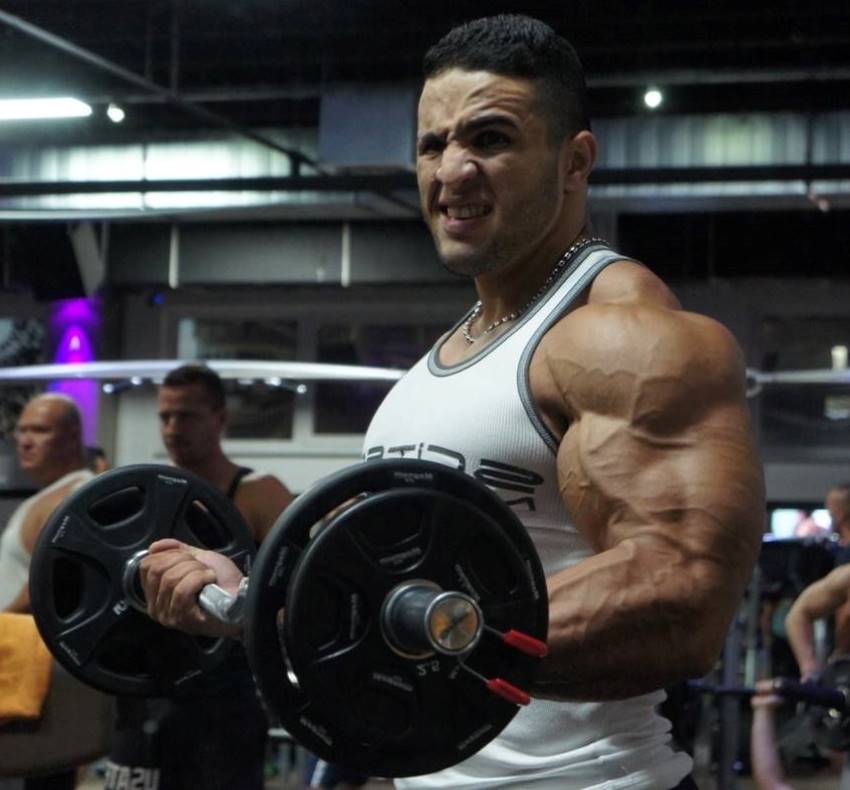 Abdelaziz Jellali doing ez-bar biceps curls in the gym, his arms looking vascular and big