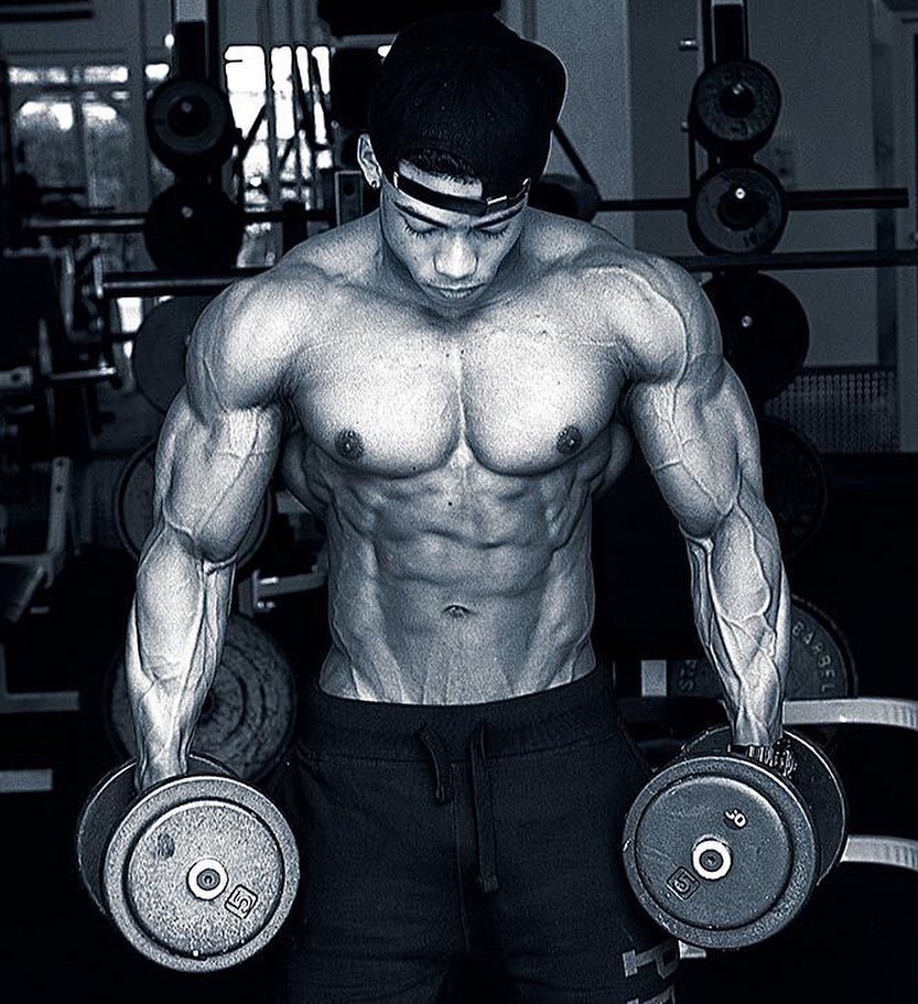 Ahmad DeGuzman holding two dumbbells and looking down, being shirtless, and looking ripped