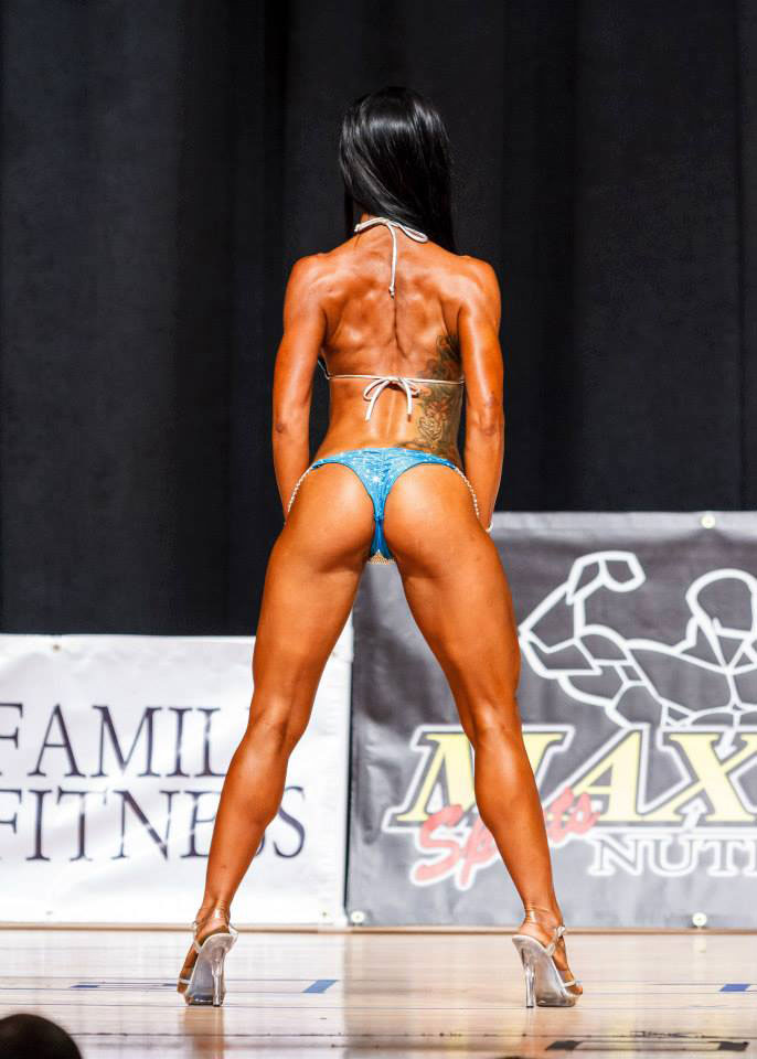 Alysia Macedo on stage at a bodybuilding competition showing off her back and glutes.