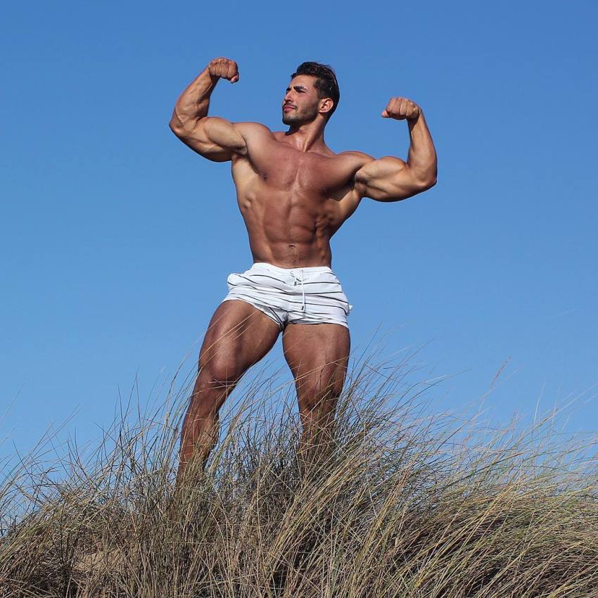 Amin Elkach doing a shirtless front double biceps flex outdoors