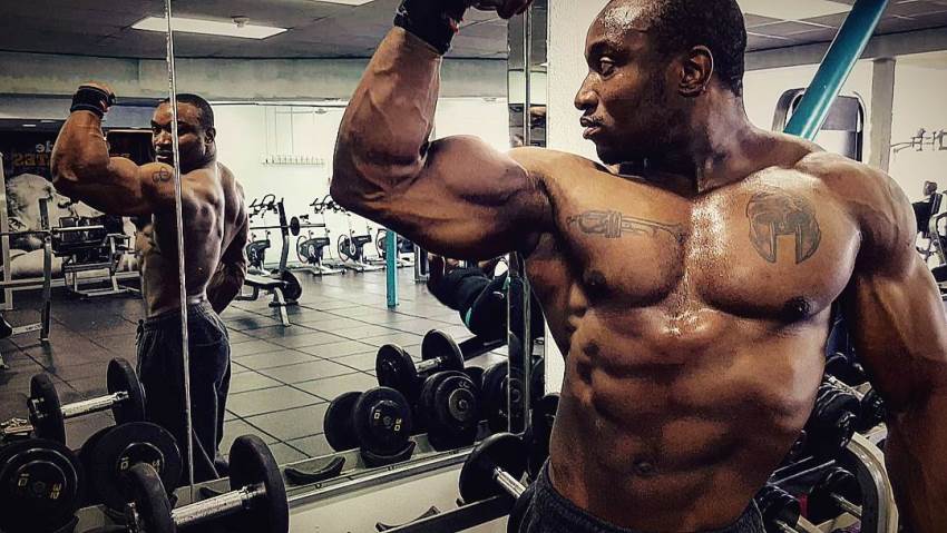 Antonio Mitchell posing shirtless in the gym, doing biceps flex in the mirror