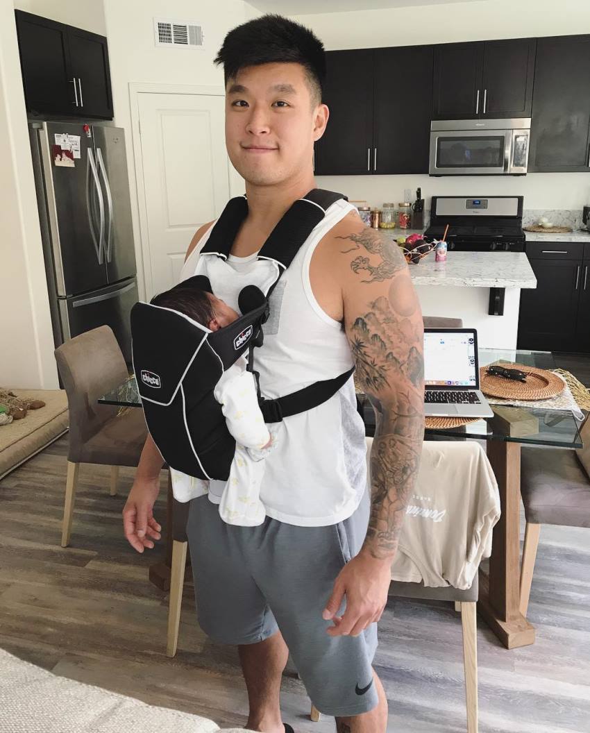 Bart Kwan carrying his child in a front facing baby carrier and smiling at the camera