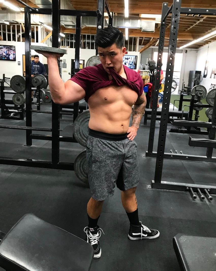 Bart Kwan holding a weight plate in the gym and showing his abs in a humorous way