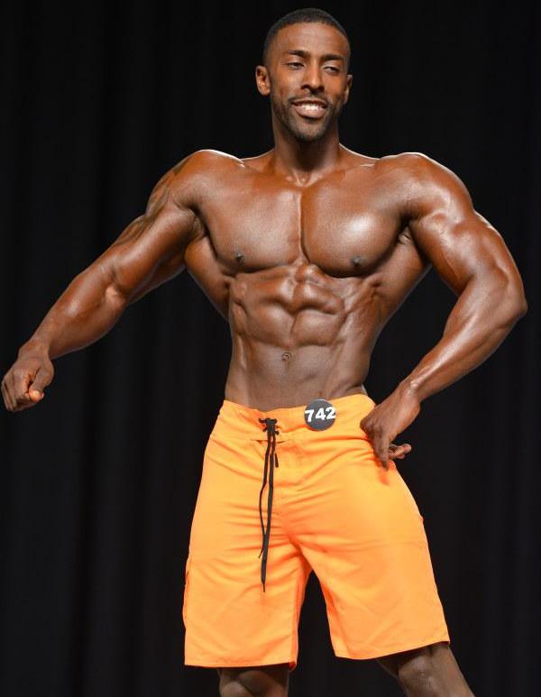 Coty Hart posing on the Men's Physique stage with a smile on his face