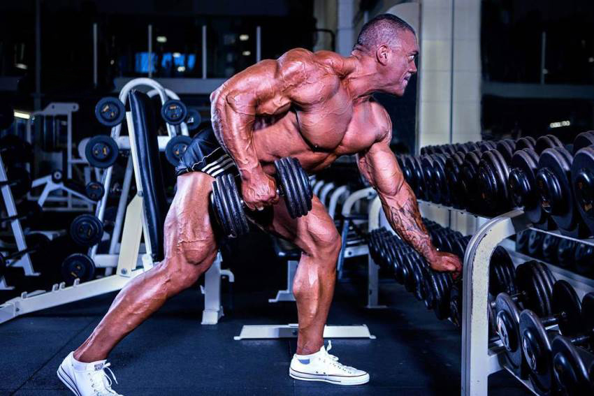 Darryn Onekawa training shirtless in the gym, doing one-arm dumbbell rows