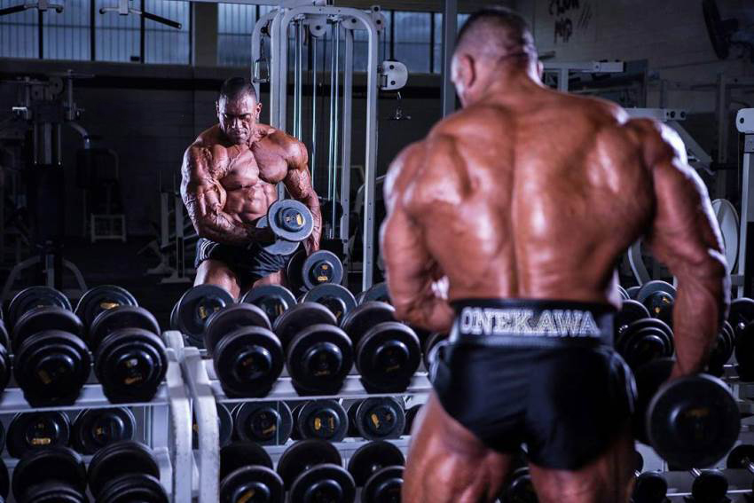 Darryn Onekawa training his arms with dumbbells in the gym shirtless, looking big and ripped