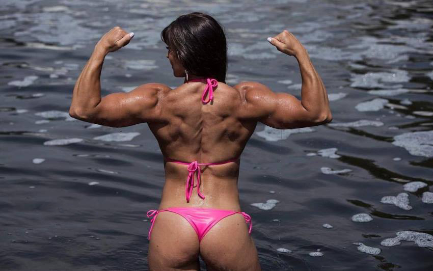 Kristen Graham doing a back double biceps pose whilst standing half-way into the sea