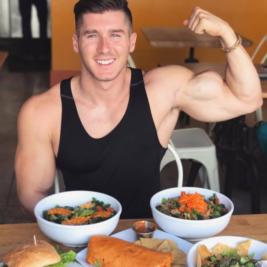 Nimai Delgado sitting with vegan food on the table and flexing his biceps