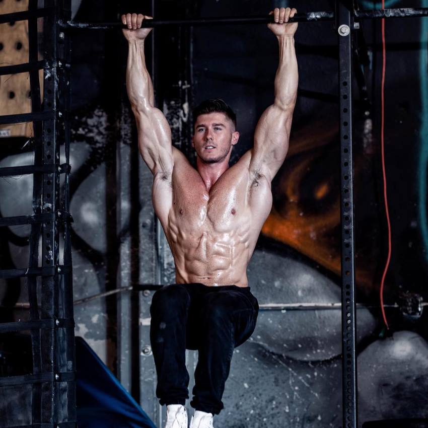 Nimai Delgado doing hanging leg raises shirtless, looking conditioned and muscular