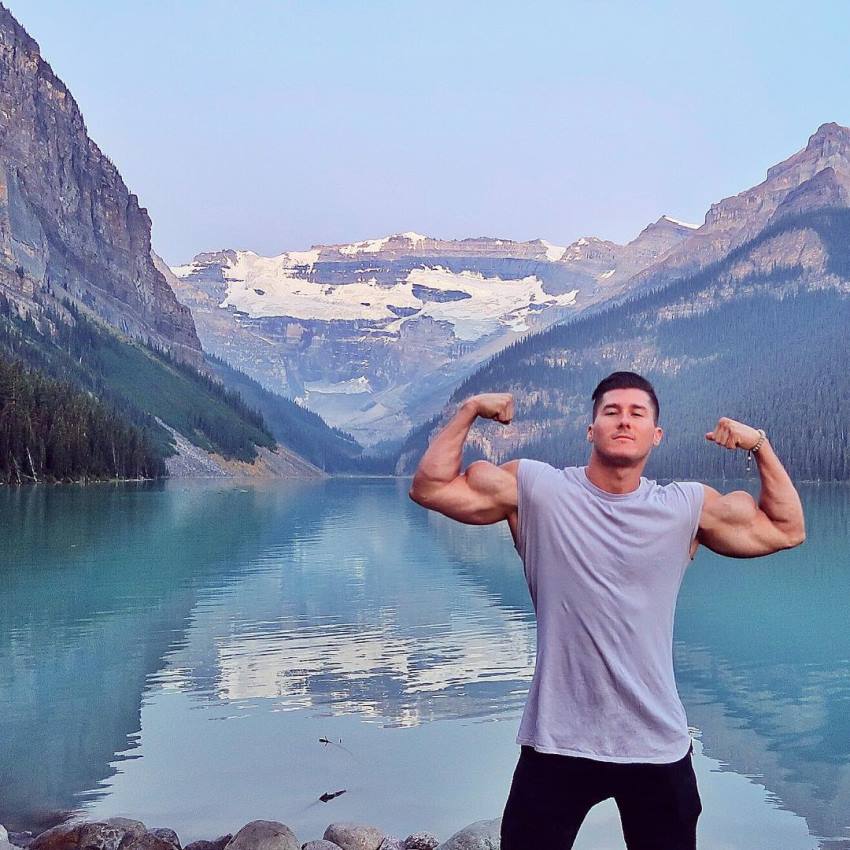Nimai Delgado flexing his arms in front of a beautiful natural scenery with lake and mountains