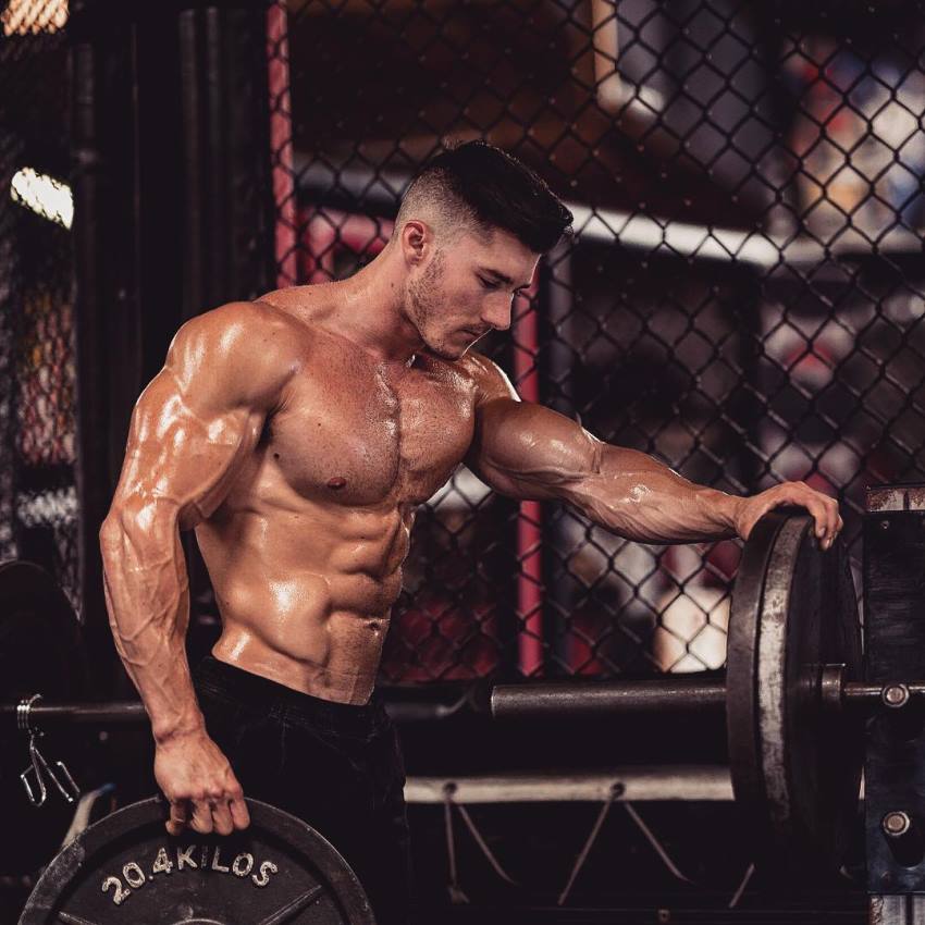 Nimai Delgado shirtless in the gym holding a weight plate with one of his muscular and ripped arms