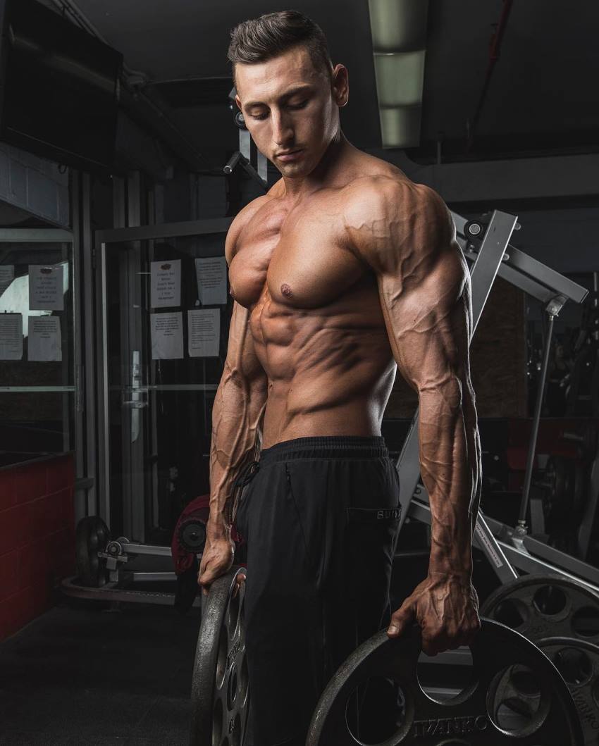 Anthony Scalza standing shirtless and holding weight plates for a photo, looking ripped and muscular