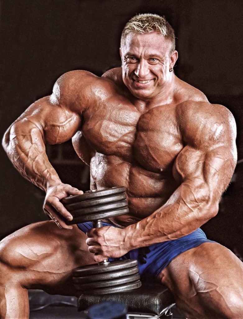 Markus Ruhl posing shirtless with a dumbbell in his hand, looking muscular and ripped