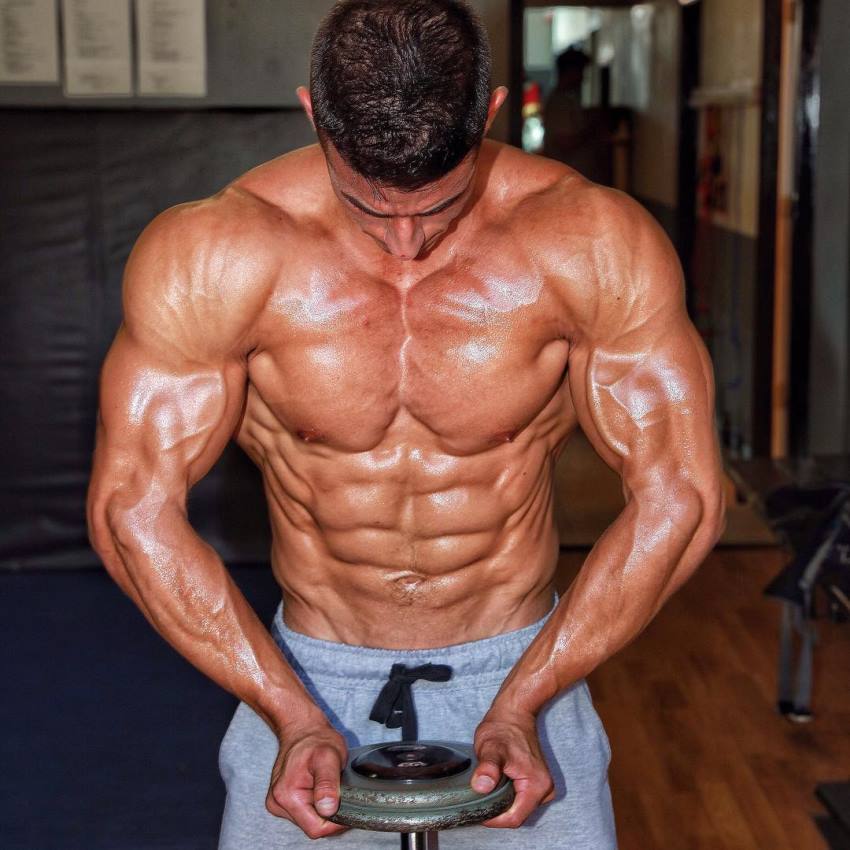 Tomas Echavarria holding a dumbbell and flexing his ripped muscles, showing hi shirtless upper body