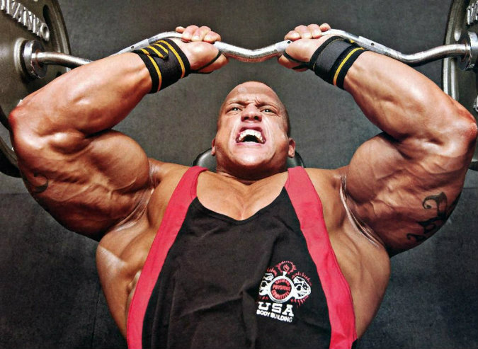 Kevin Jordan doing heavy skullcrushers with his massive arms, having a pained expression on his face while doing the exercise