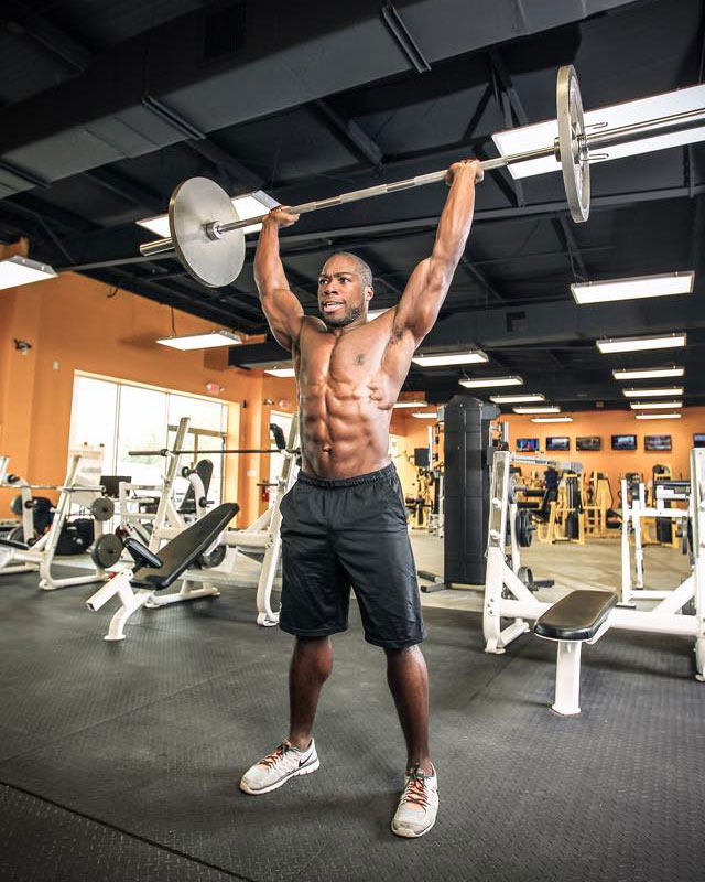 Kizzito Ejam performing an overhead press with a barbell.