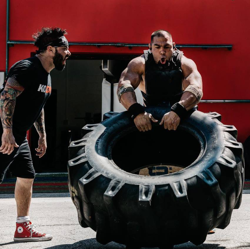 Tony Sentmanat flipping a big heavy tire while another man is motivating him by yelling at him