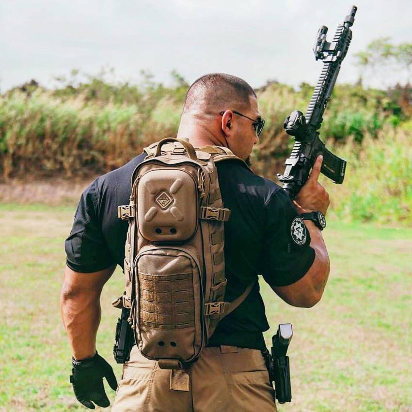 Tony Sentmanat wearing a combat-equipment backback along with a rifle in his arm, looking big and muscular