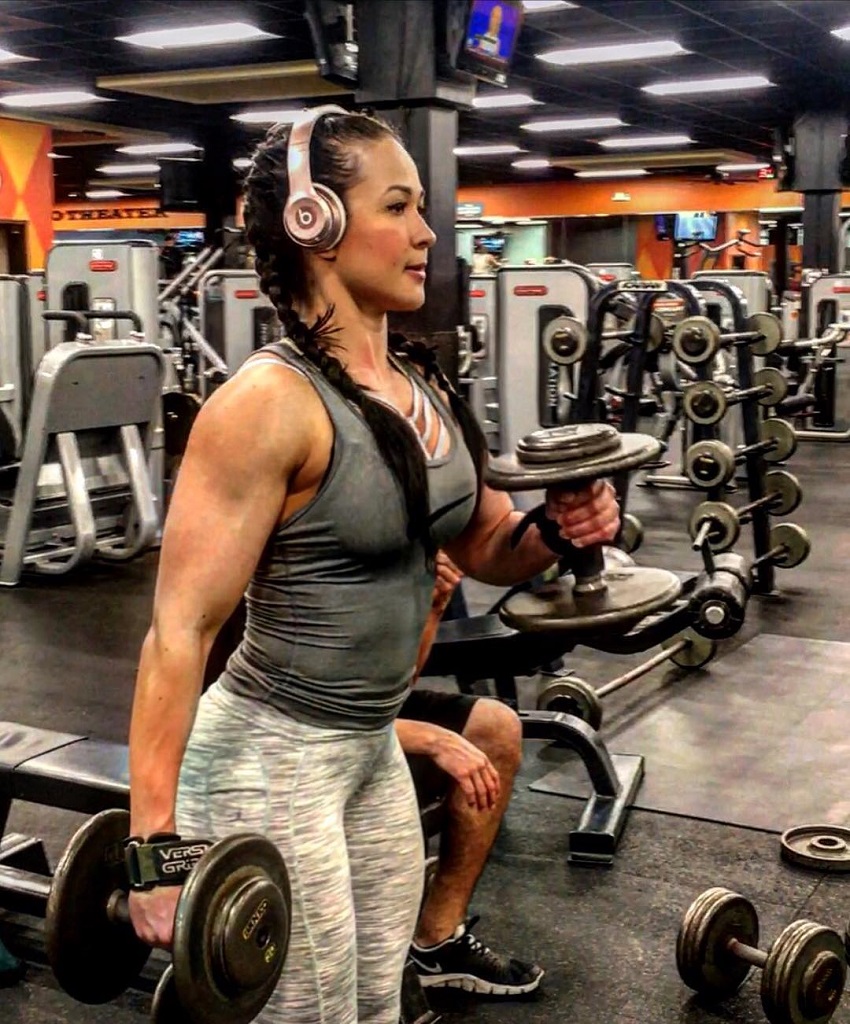 Alexis Mariah Avina doing dumbbell curls in the gym with headphones on, looking fit and lean