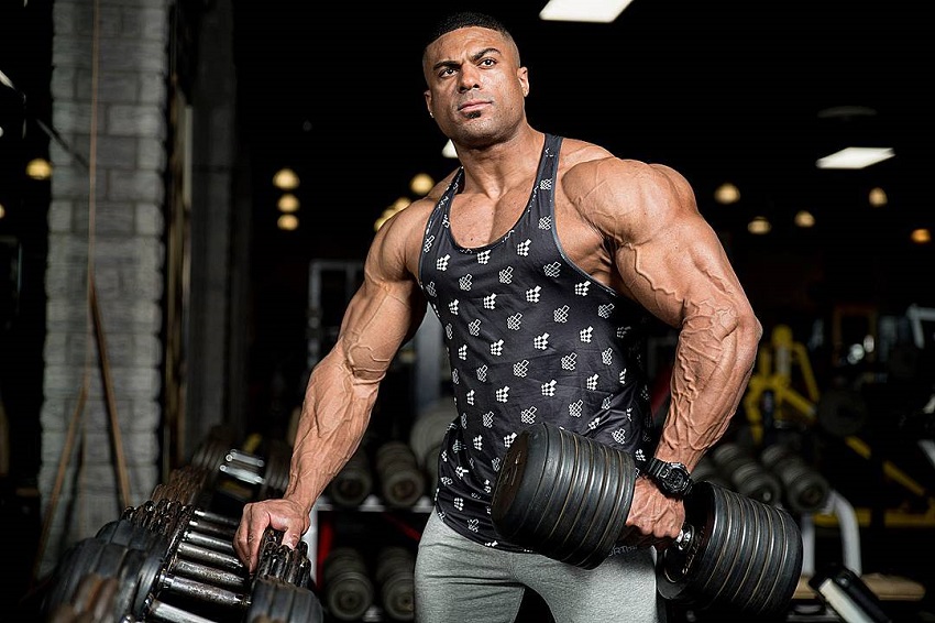 Henri-Pierre Ano holding a dumbbell in one of his hands and leaning against a dumbbell rack with the other hand, looking ripped and muscular