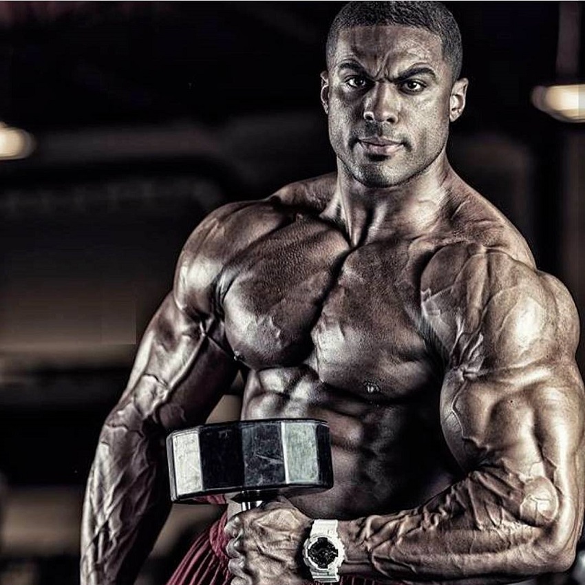 Henri-Pierre Ano lifting a dumbbell while looking at the camera, looking ripped and lean