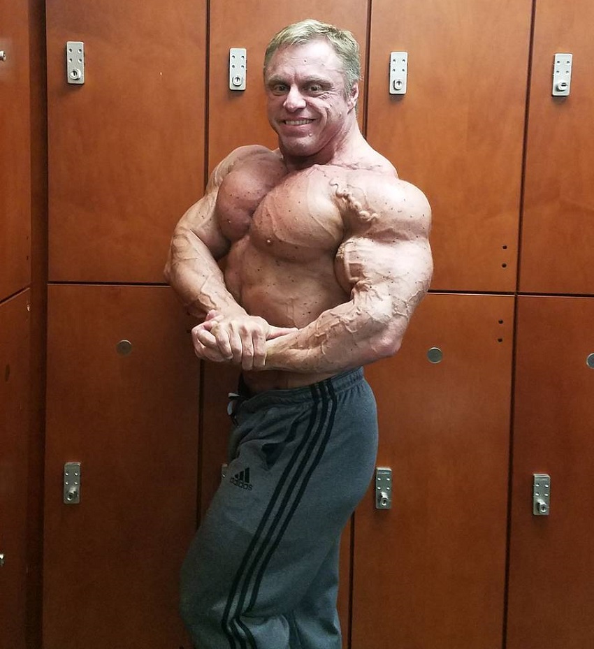 John Meadows doing a shirtless side chest pose in a gym locker room