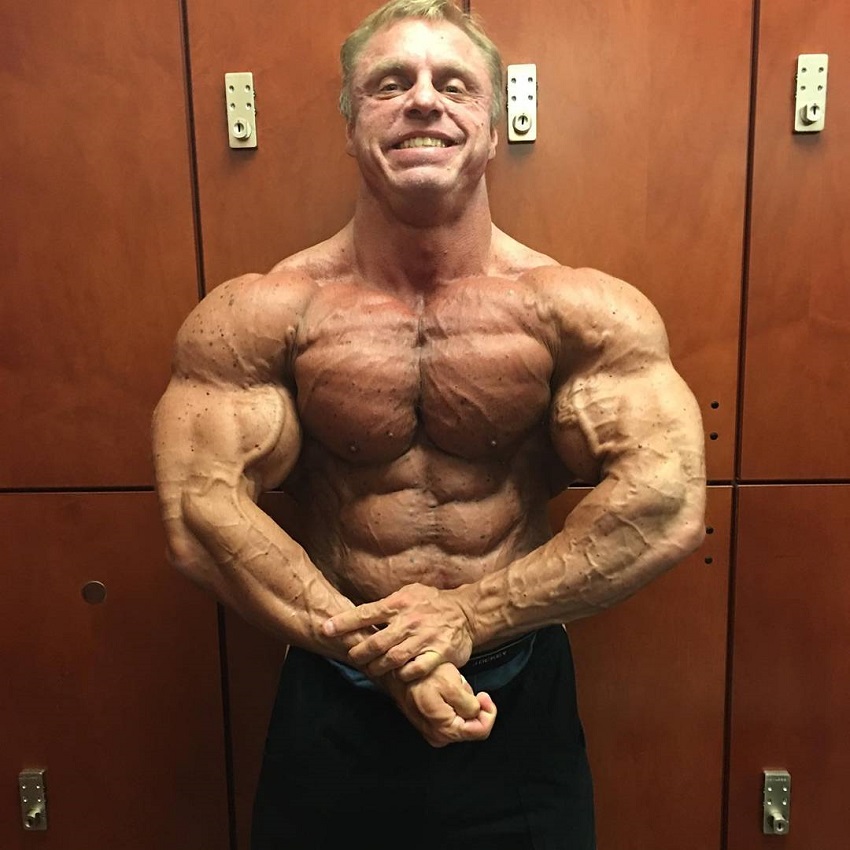 John Meadows doing a shirtless most muscular pose in a gym locker room