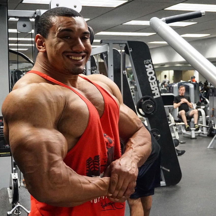 Larry Wheels doing a side chest pose in a gym wearing a red tank top