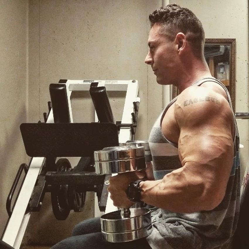 Micah Lacerte sitting on a bench with dumbbells in his hands looking ripped and muscular