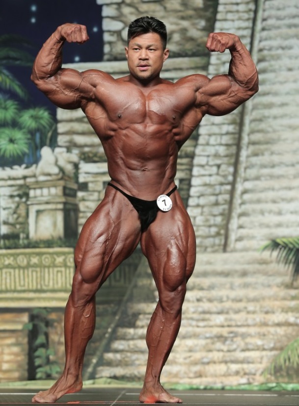 An Nguyen doing a front double biceps pose on a bodybuilding stage