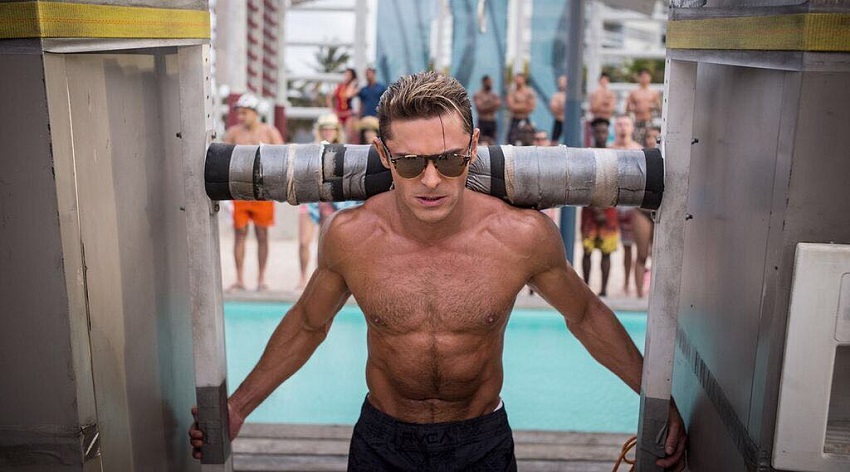 Zac Efron lifting a construct in front of numerous people that stand by the pool, looking ripped and fit