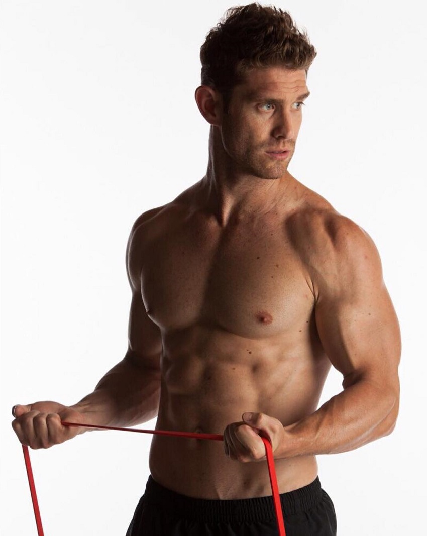 CJ Koegel posing shirtless with resistance bands in his arms, looking aesthetic and lean