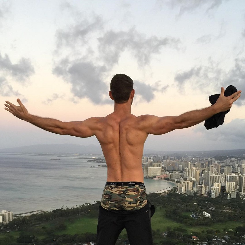 CJ Koegel overlooking a big city and nature with his arms wide open, looking muscular and lean